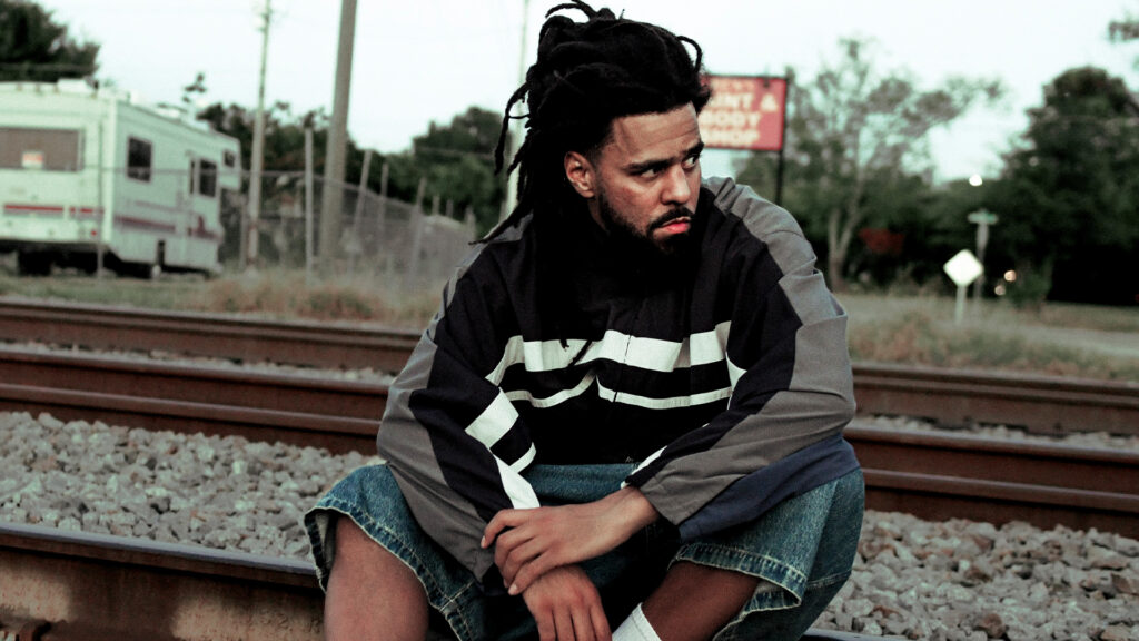 Rapper J Cole sitting on train tracks, staring into the distance.